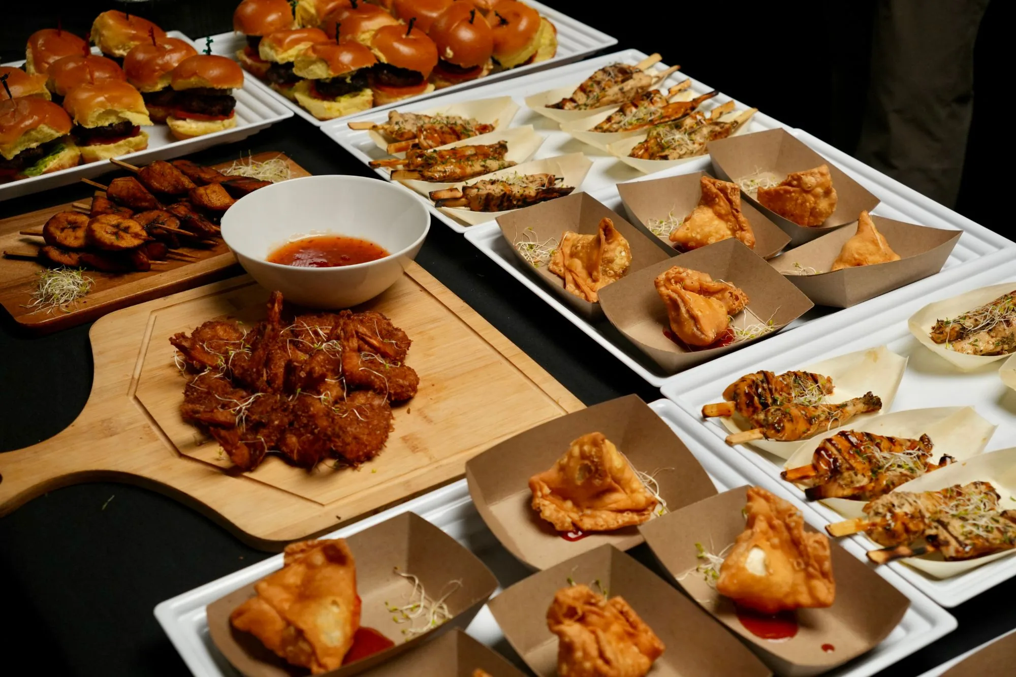 A catering spread of sliders, chicken skewers, samosas and coconut shrimp on wooden boards and white platters